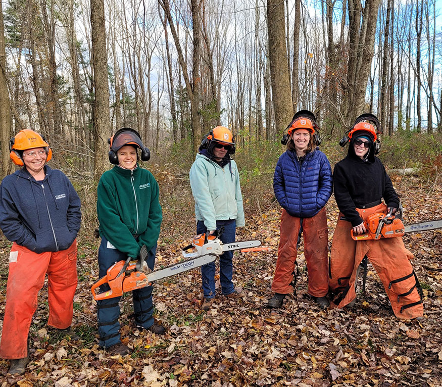 Five women in chainsaw gear posing for a photo in the woods holding their chainsaws. 