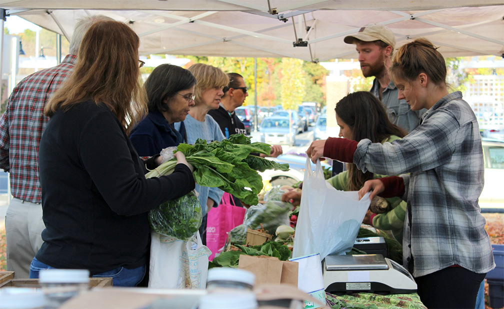 group of people under a tent buying vegetables at a farmers market.